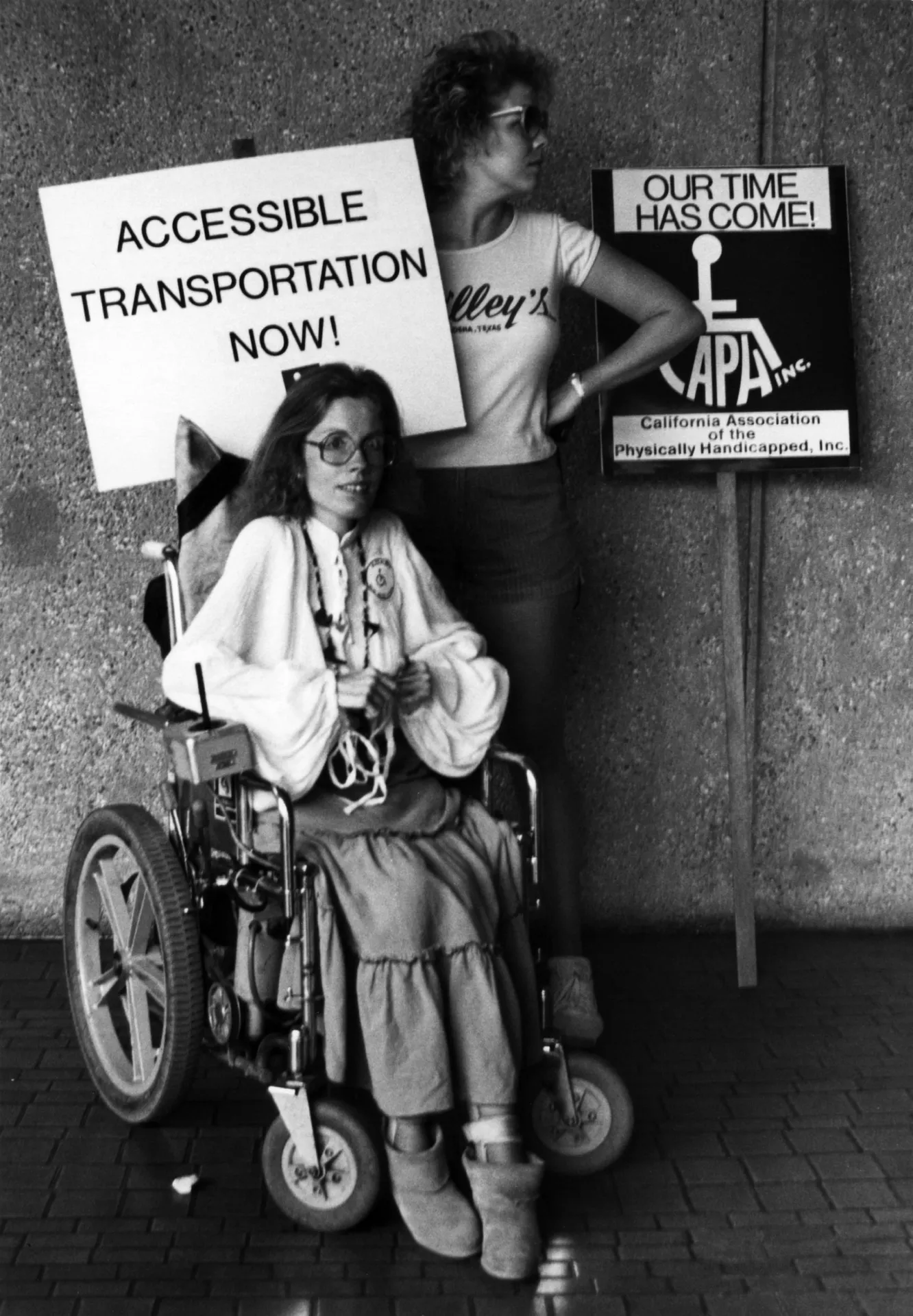 Diane Coleman in Los Angeles sitting in her motorized wheelchair with protest sign which reads “Accessible Transportation Now”.