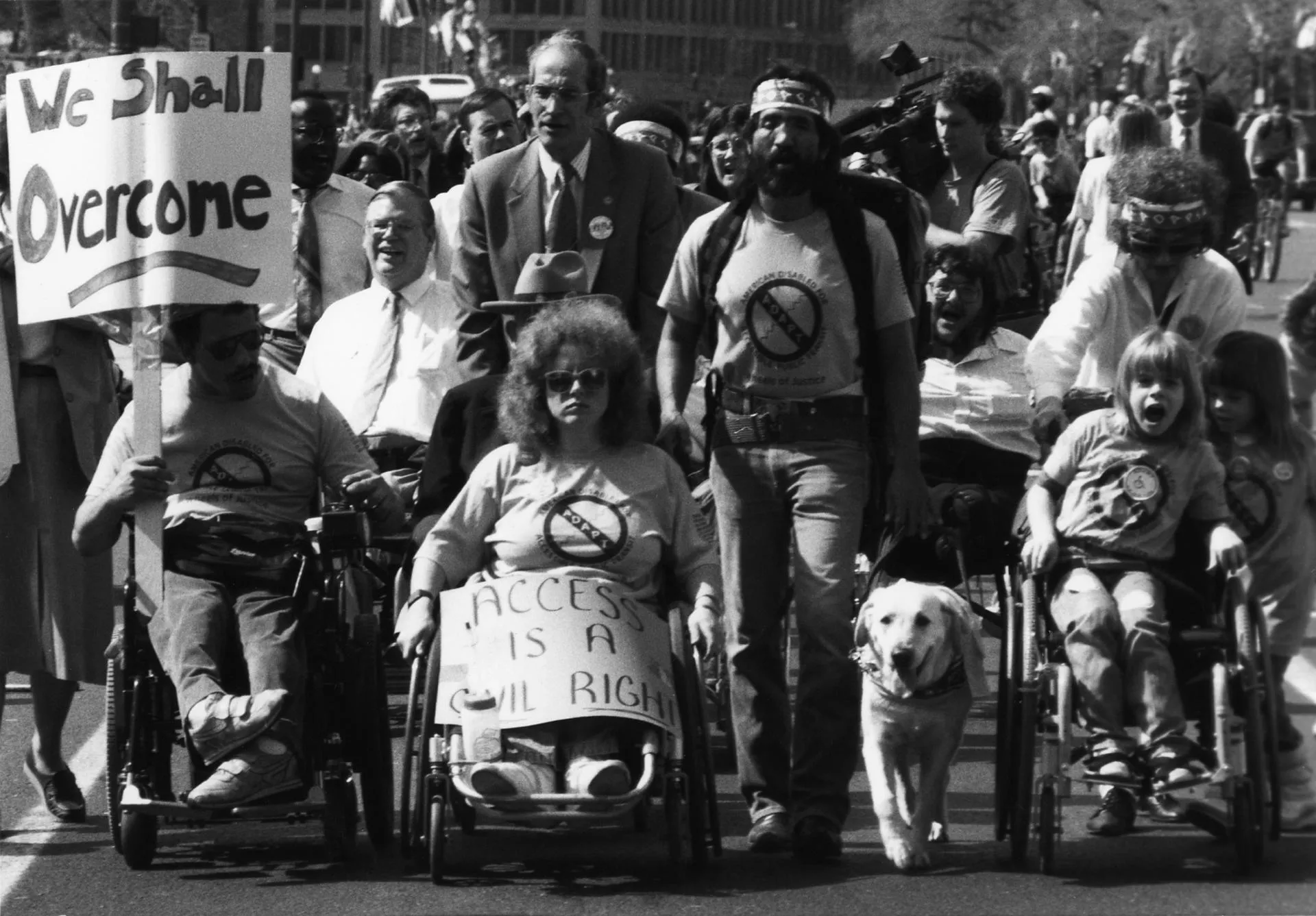 Protest sign that reads We Shall Overcome! March of adult and children protesting. Protestors are in wheelchairs, using service animals and walking.