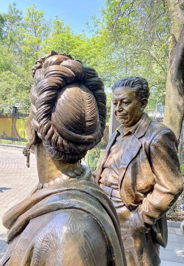 Two statues are captured in a tree-filled plaza.  One statute is a man white short wavy hair in a blazer looking down.  The woman statute’s picture is taken from behind.  Her braided hair is wrapped around her head, she is wearing earrings, a large beaded necklace, and a shawl on her shoulders.