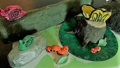 This is a photograph of a sculptural work of art made of mixed-media papier-mache and depicts a still-life of small creatures dwelling in a natural habitat; there are two brightly colored snails, two worms, and one butterfly perched on top of natural elements such as stone, wood, grass, and leaves.
