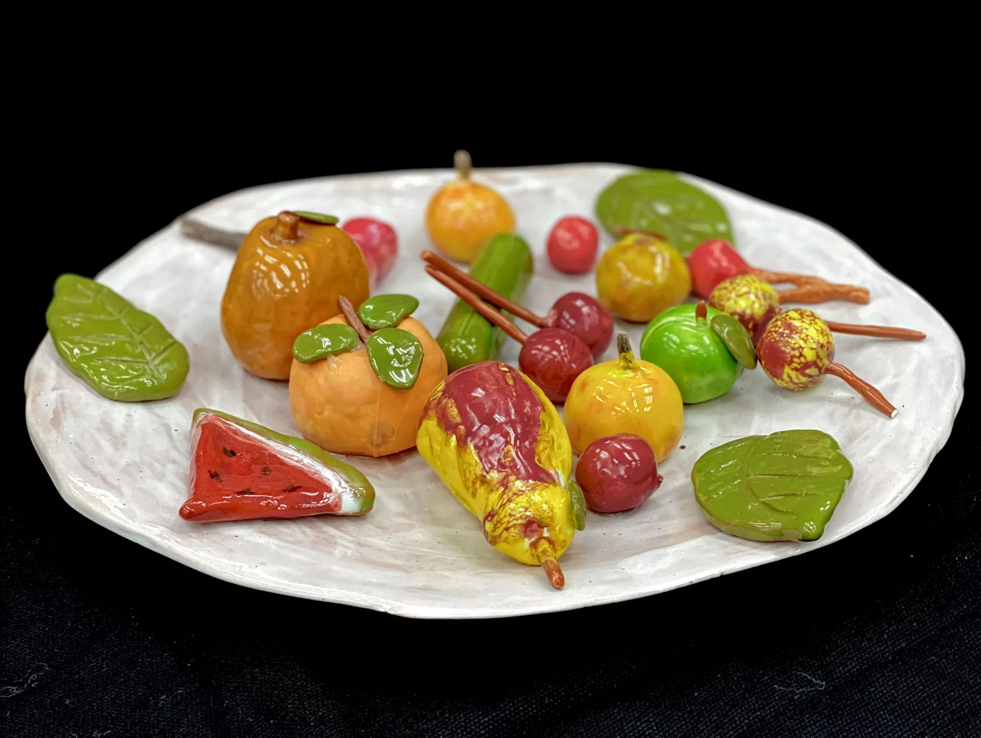 Anet Shahbandari Fruit plate, 2020 The two photographs depict a ceramic sculpture of a plate of intensely colored fruit: citrus fruit, cherries, a slice of watermelon, and decorative green leaves.On photograph is taken from directly above and the other forward tilted angle.  Ceramic, 12” x 12” x 3"
