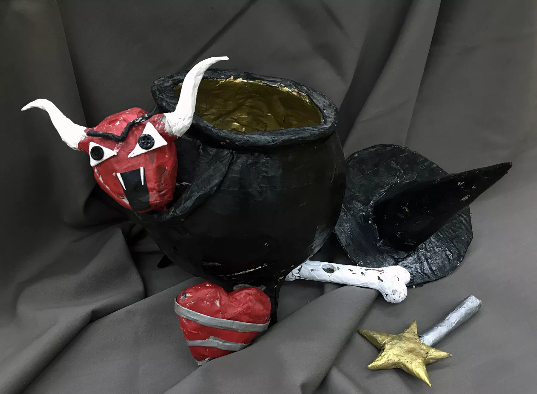 Cristian Valenzuela The Cauldron of Evil, 2022 The paper-mache form was created in an "Art & Reading" class and inspired by the strange creatures from the original Grimm's Brothers fairy tales. The white horned head of a sinister red faced bull is fixed to a black cauldron. Surrounding the cauldron is a red heart wrapped in silver stripes, a silver wand tipped with a golden star, a white bone, and a witches pointed hat.  Paper-Mache, 19” x 19” x 14”