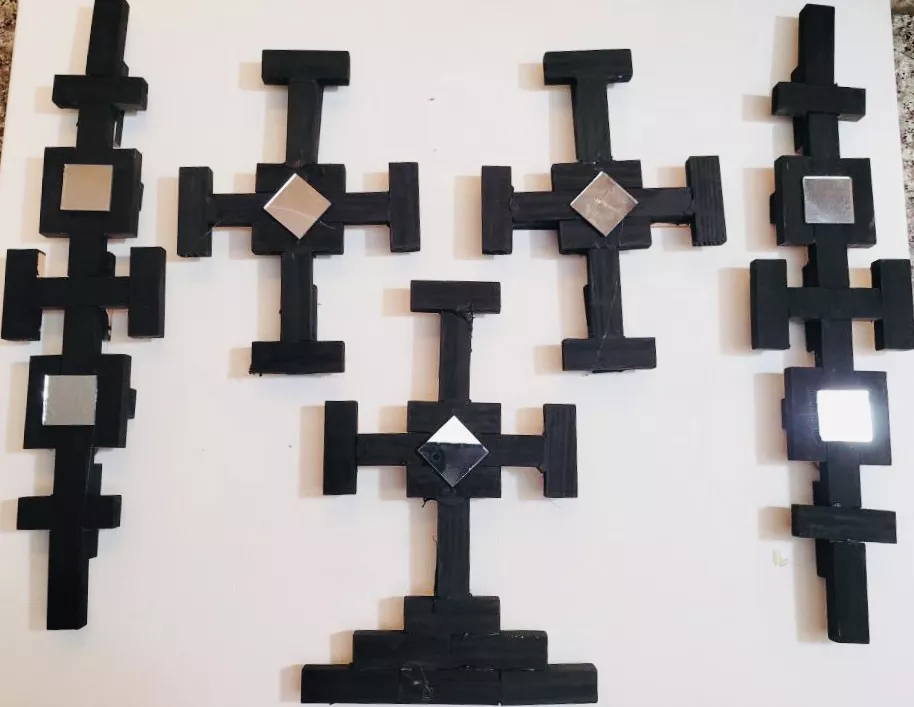 Jesus Herrera Tribal Jenga Vibes, 2022 Small black geometric wooden forms made up of rectangle jenga pieces. Two boarding forms are each a single length made up of rectangles and two squares. There are three t-shaped forms, one of which has three layers at the bottom. These forms have a singular center square. All the squares have reflective mirror pieces.  Upcycled wooden "Jenga" game pieces, glue, mirrored tiles, 18" x 24"