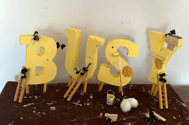 James Sanchez Busy as a Bee, 2022 A team of small bees are finishing the construction of the letters, B, U, S, Y (BUSY). Bees on work ladders are painting the letters yellow using paint cans, bushes and paint rollers. Tools and paint supplies lie on the brown ground. The letters S and Y’s construction is almost complete and the honeycomb structure underneath can still be seen.  Foam, ground coffee, wooden dowels, toothpicks, hot glue, bubble wrap, pom poms, cut plastic and miniature tools, 9" x 22" 9"