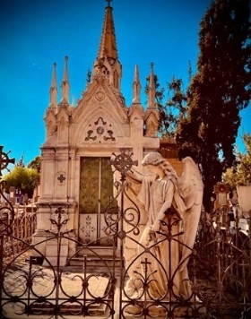 A photograph of a gated mausoleum in Mexico, designed as a small steepled church. A statue of a full-sized classically winged angel looks down towards a grave.