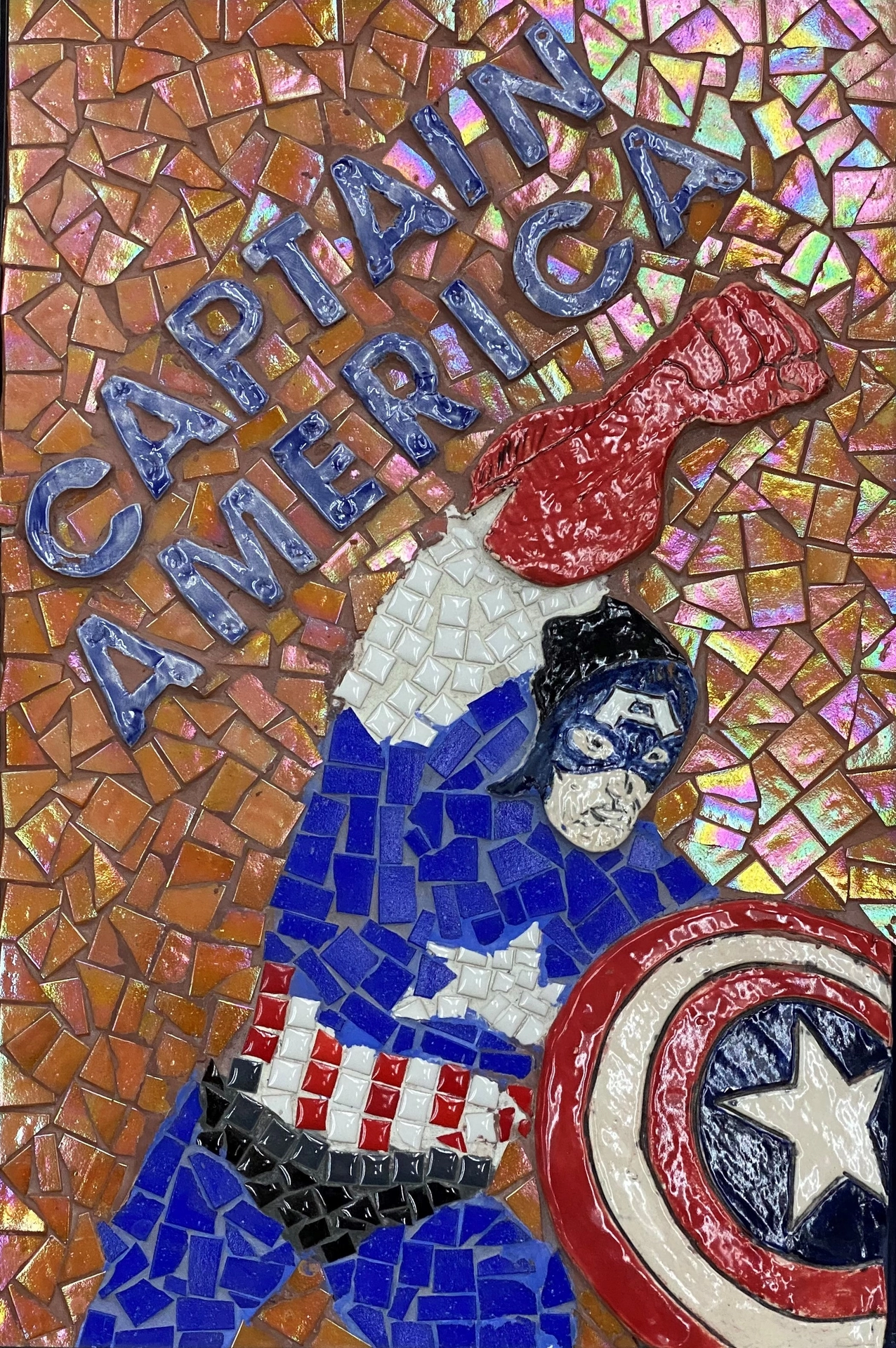 This handmade mosaic/ceramic piece in vertical rectangular format is framed in black. The classic American comic book hero Captain America is wearing his iconic red, white, and blue costume. His arm is outstretched up and out in a display of his power. The background is made up of broken orange pieces of ceramic tile.